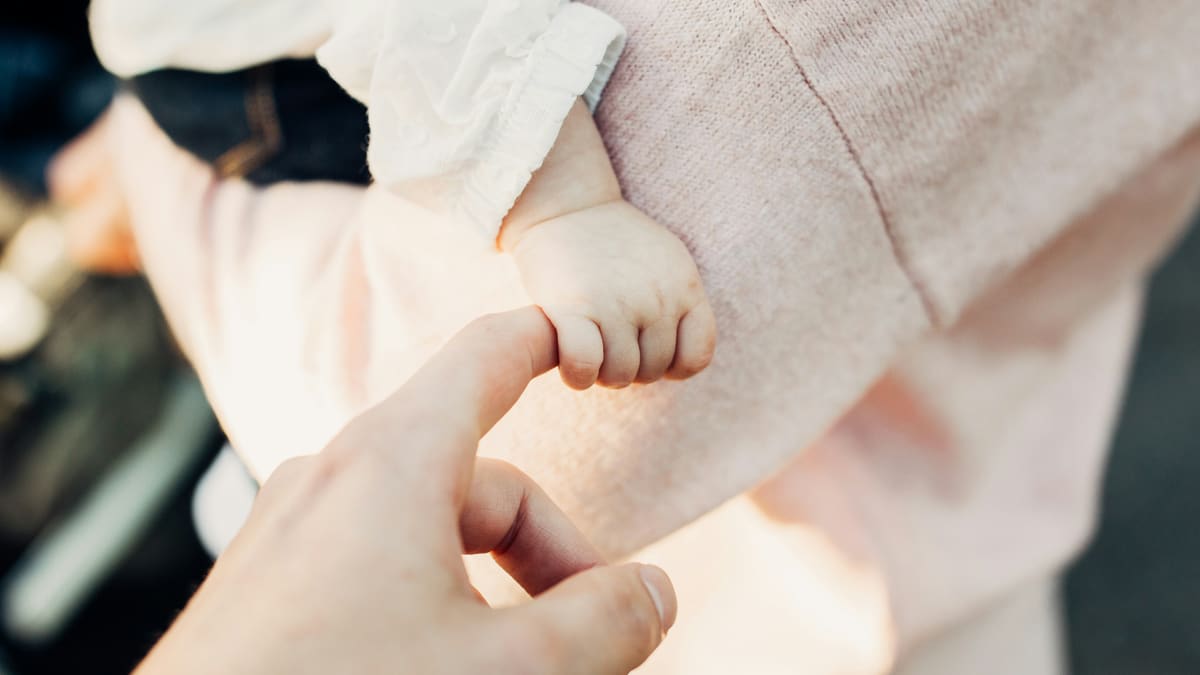 A mãozinha de um bebê agarrada ao dedo de um adulto, com roupas em tons de rosa desfocadas ao fundo