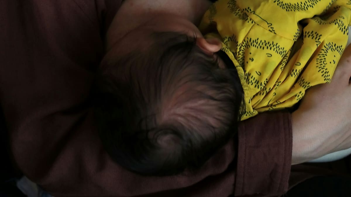 A cabeça de um recém-nascido vista de cima, no colo de quem o amamenta, ao lado de uma roupinha amarela estampada